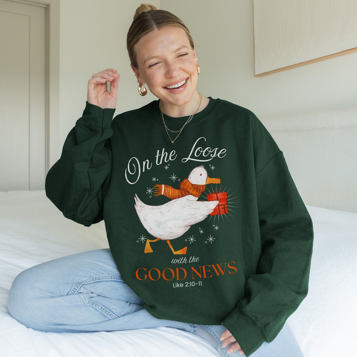 Woman wearing a green sweatshirt with a goose graphic and text, sitting on a bed.