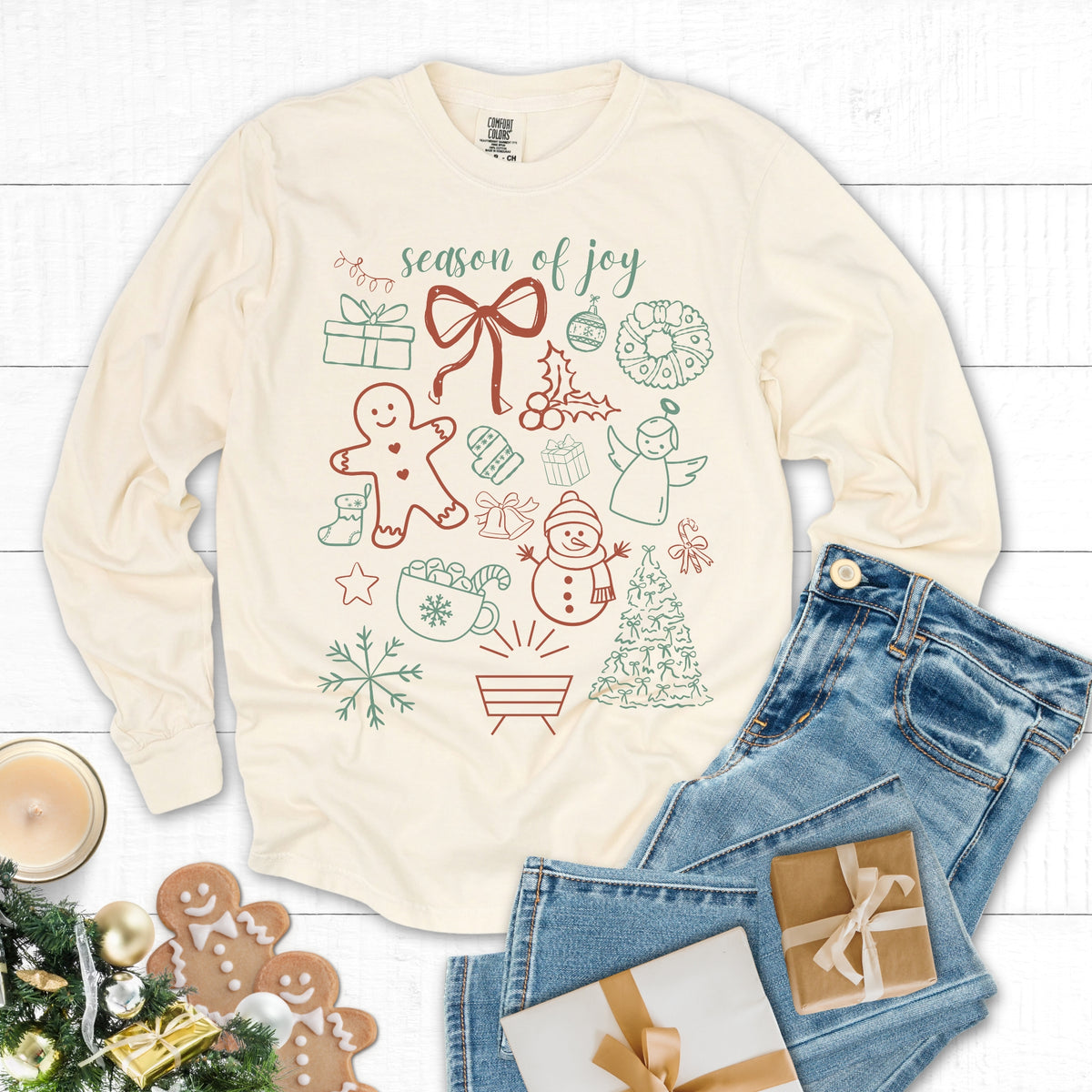 Beige long-sleeve shirt with Christmas-themed graphics on a white wooden surface with jeans and gift boxes.