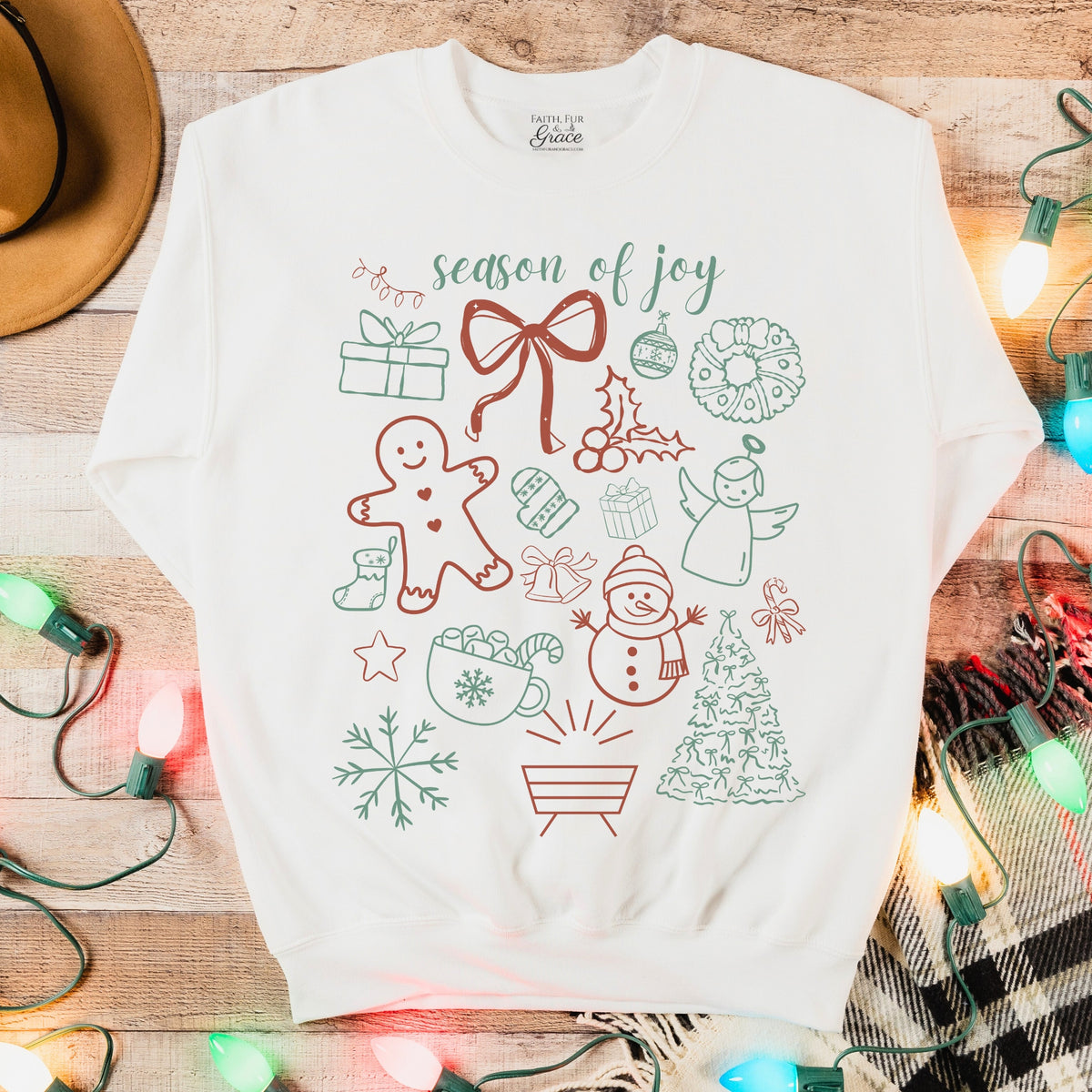 White sweatshirt with Christmas-themed graphics on a wooden surface with lights and plaid fabric.