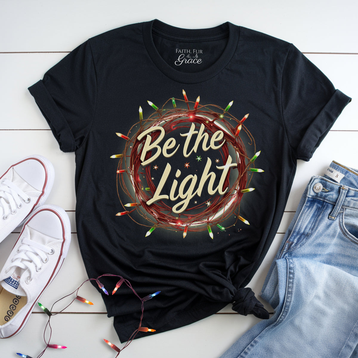 Black t-shirt with 'Be the Light' design on a white wooden surface with sneakers and jeans.