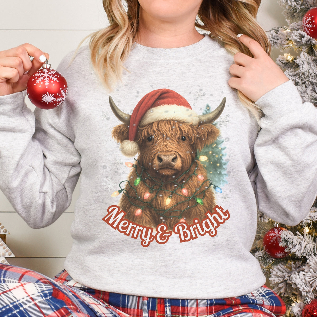 Person wearing a Christmas-themed sweatshirt with a highland cow and ornaments, holding a red ornament.
