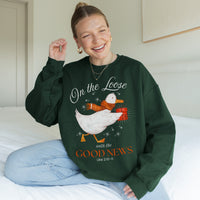 Woman wearing a green sweatshirt with a goose graphic and text, sitting on a bed.