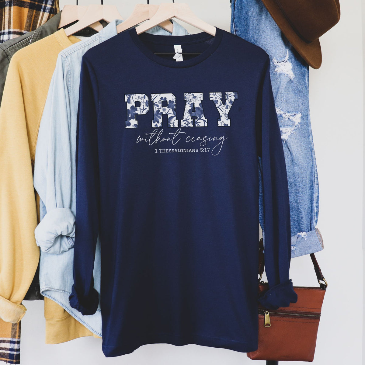 Navy long-sleeve shirt with 'PRAY without ceasing' text on a rack with clothes and a handbag.