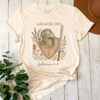 Beige t-shirt with armor graphic and text on a wooden surface with plants.