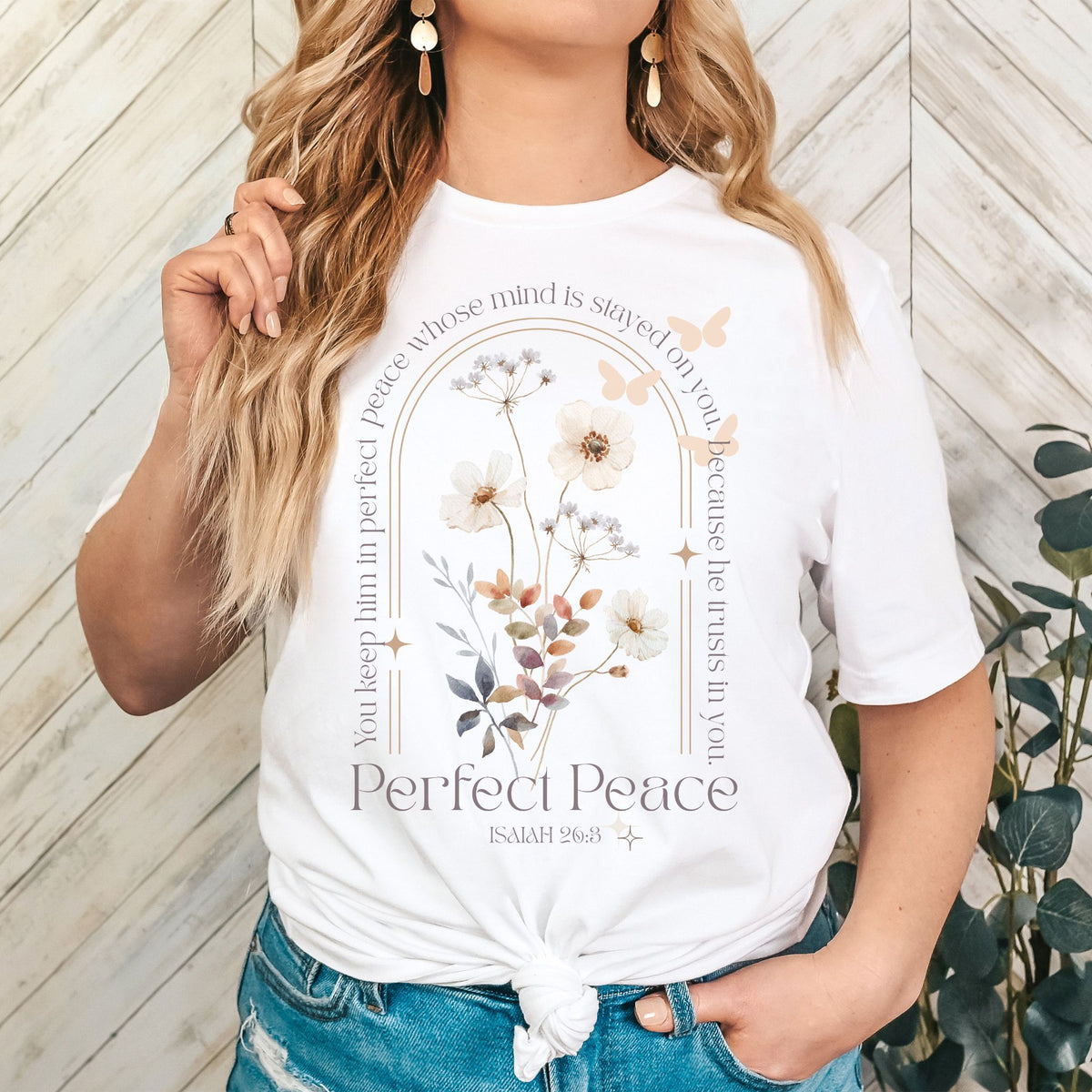 Person wearing a white t-shirt with floral design and text, standing against a wooden wall.