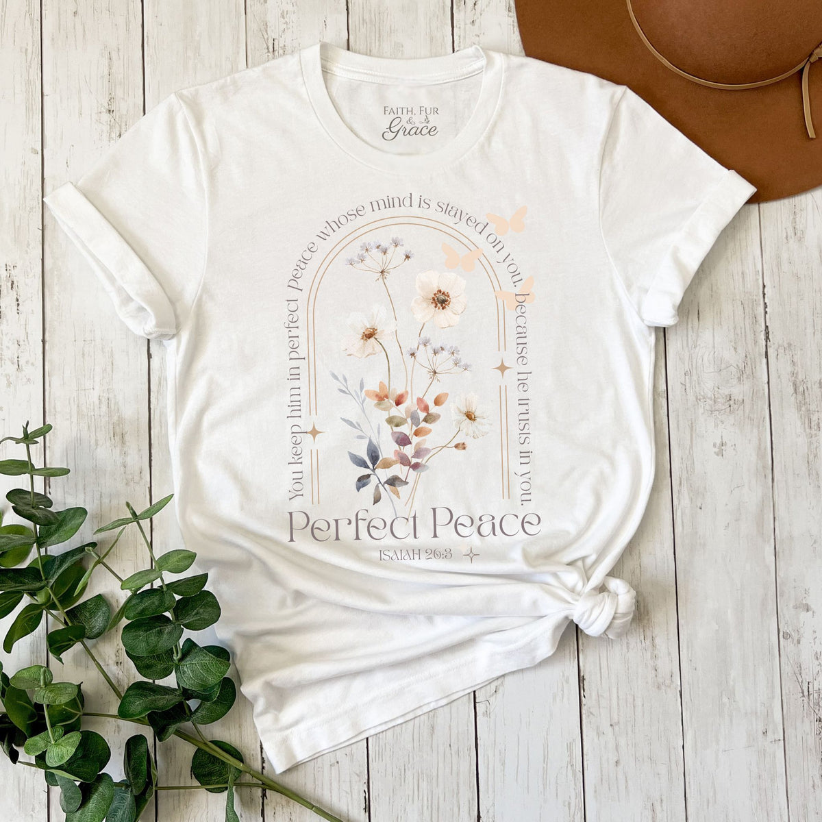 White t-shirt with floral design and text on a wooden surface with a plant and brown hat.