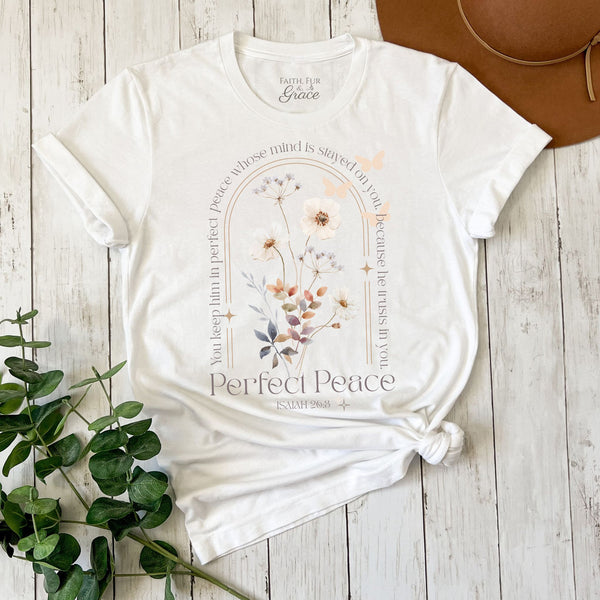 White t-shirt with floral design and text on a wooden surface with a plant and brown hat.