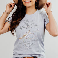 Woman wearing a t-shirt with a bird graphic and text, holding a hat.