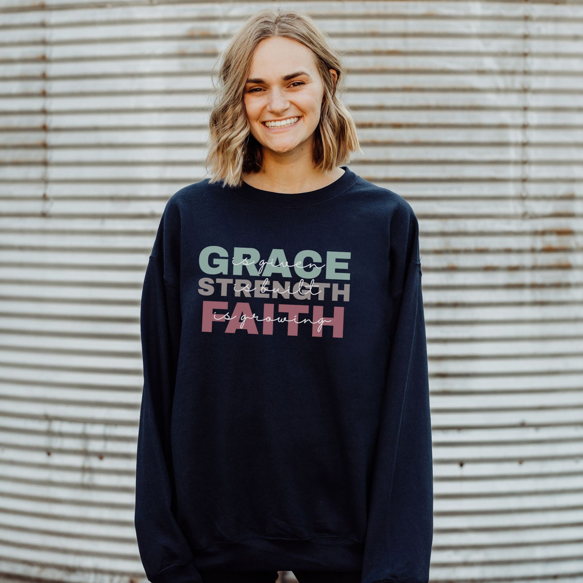 Person wearing a navy sweatshirt with 'GRACE STRENGTH FAITH' text against a corrugated metal background
