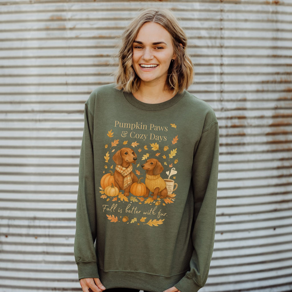Woman wearing a green sweatshirt with 'Pumpkin Paws & Cozy Days' design against a corrugated metal background.