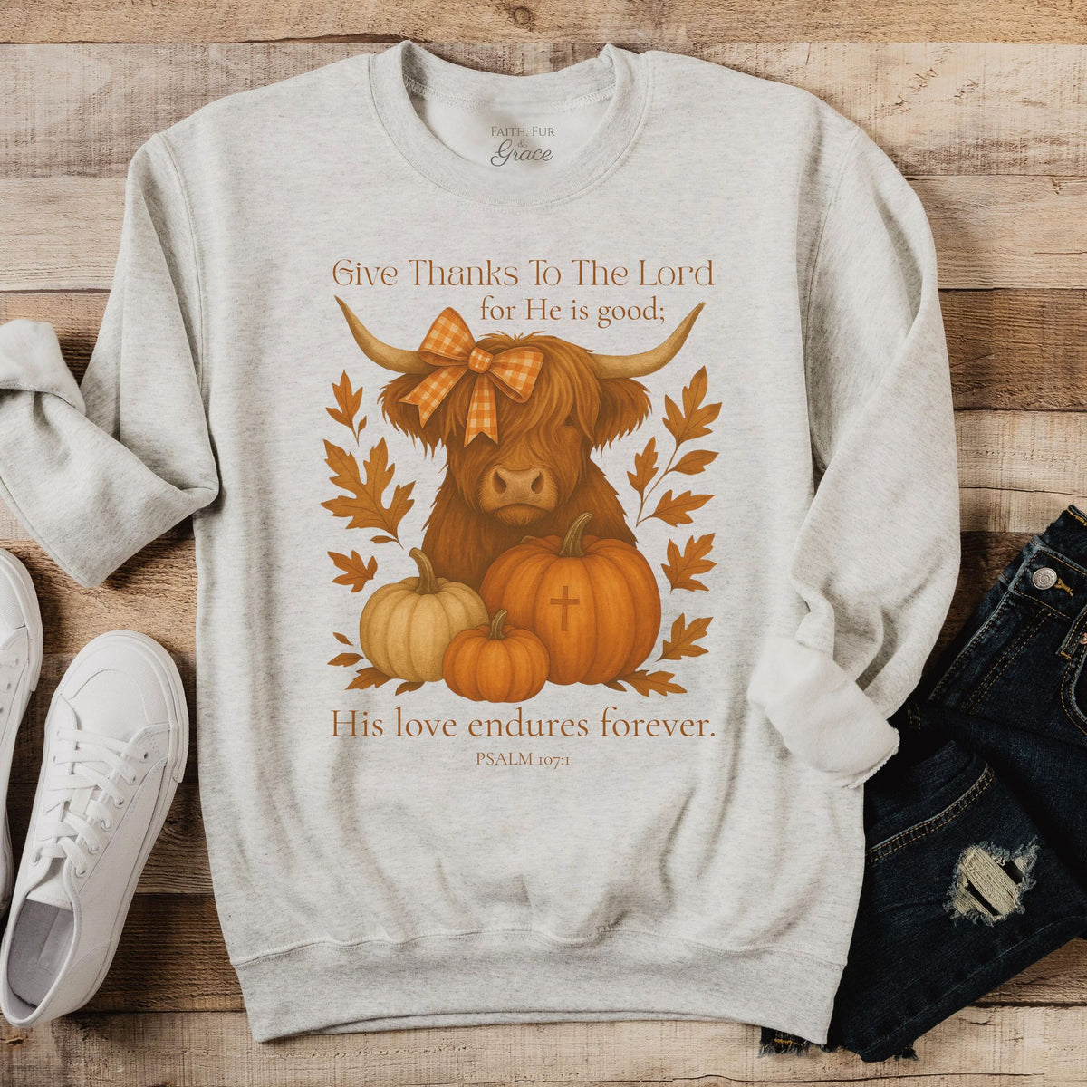 Gray sweatshirt with a cow and pumpkins design on a wooden surface with shoes and jeans.