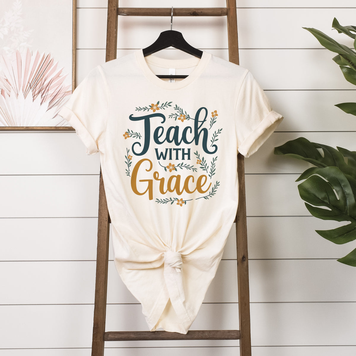 White t-shirt with 'Teach with Grace' text on a wooden ladder against a white wall.
