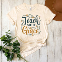 Beige t-shirt with 'Teach with Grace' text on a wooden surface with a plant and hat.
