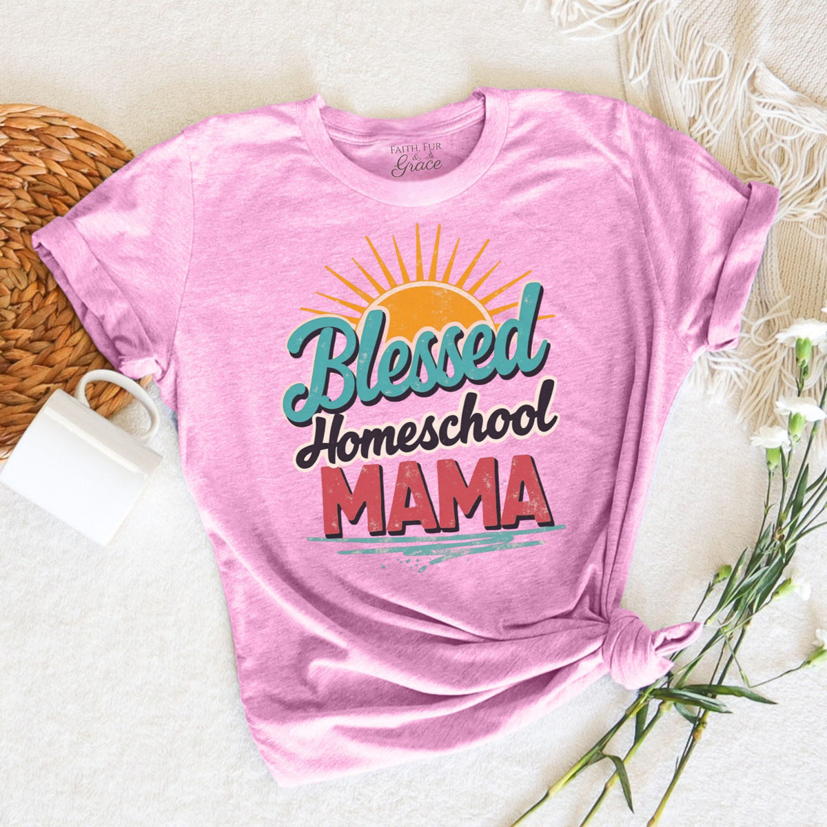 Pink t-shirt with 'Blessed Homeschool Mama' text on a white surface with a woven mat and flowers.