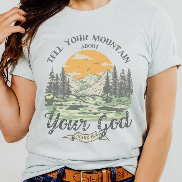Light blue t-shirt with nature graphic and text 'Tell Your Mountain About Your God' worn by a person.