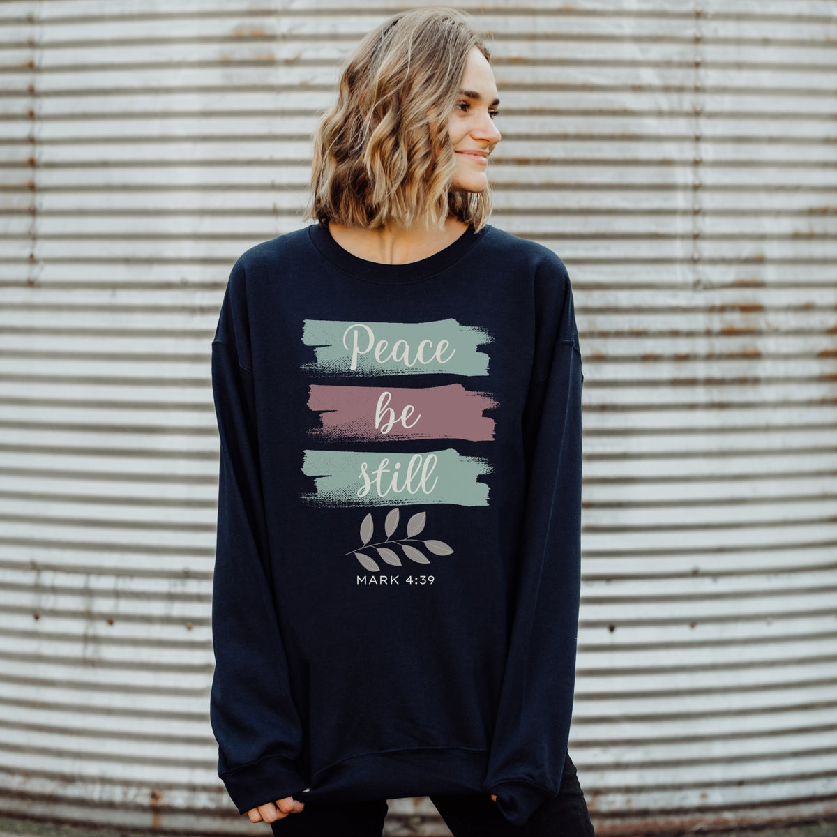 Woman wearing a navy sweatshirt with 'Peace be still' text against a corrugated metal background