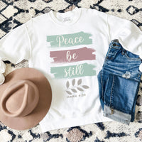 White sweatshirt with 'Peace be still' text, jeans, and hat on a patterned surface