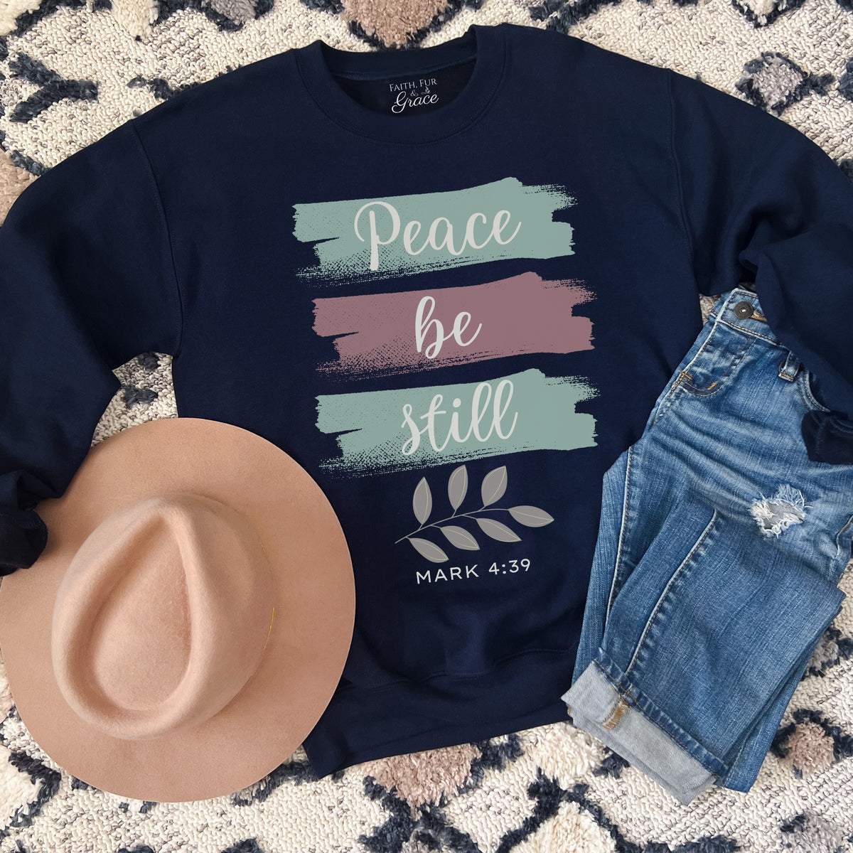 Navy sweatshirt with 'Peace be still' text, hat, and jeans on a patterned surface