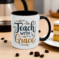 White mug with black interior and handle, featuring 'Teach with Grace' text on a wooden table with coffee beans and a slice of cake.