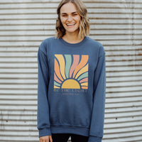 Person wearing a blue sweatshirt with a colorful sun design and text against a corrugated metal background.