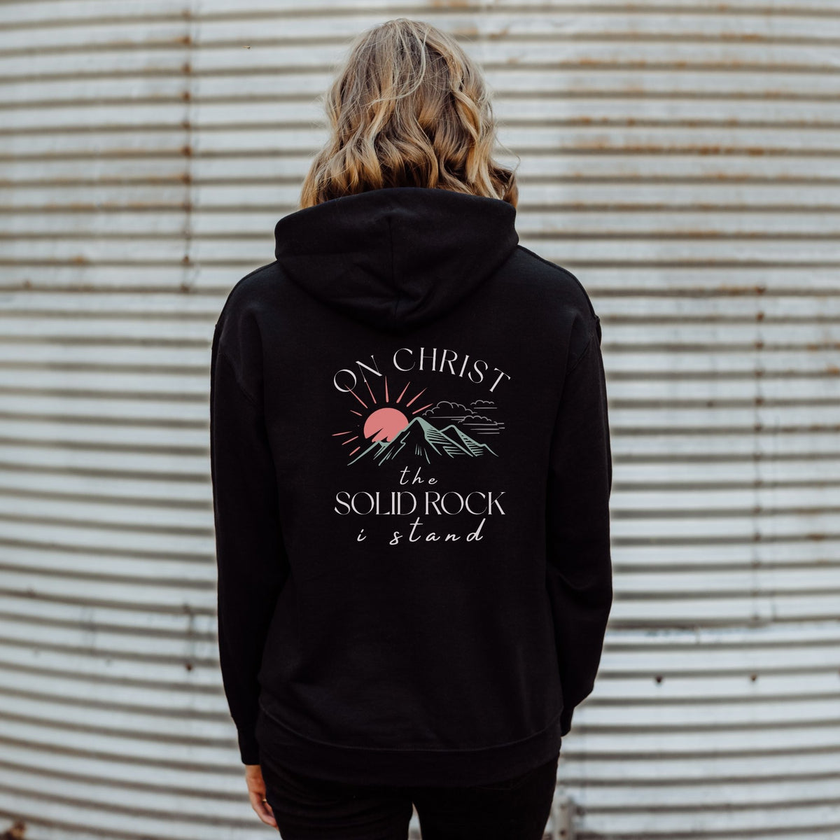 Person wearing a black hoodie with a design and text on the back against a corrugated metal wall.