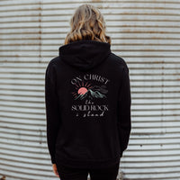 Person wearing a black hoodie with a design and text on the back against a corrugated metal wall.