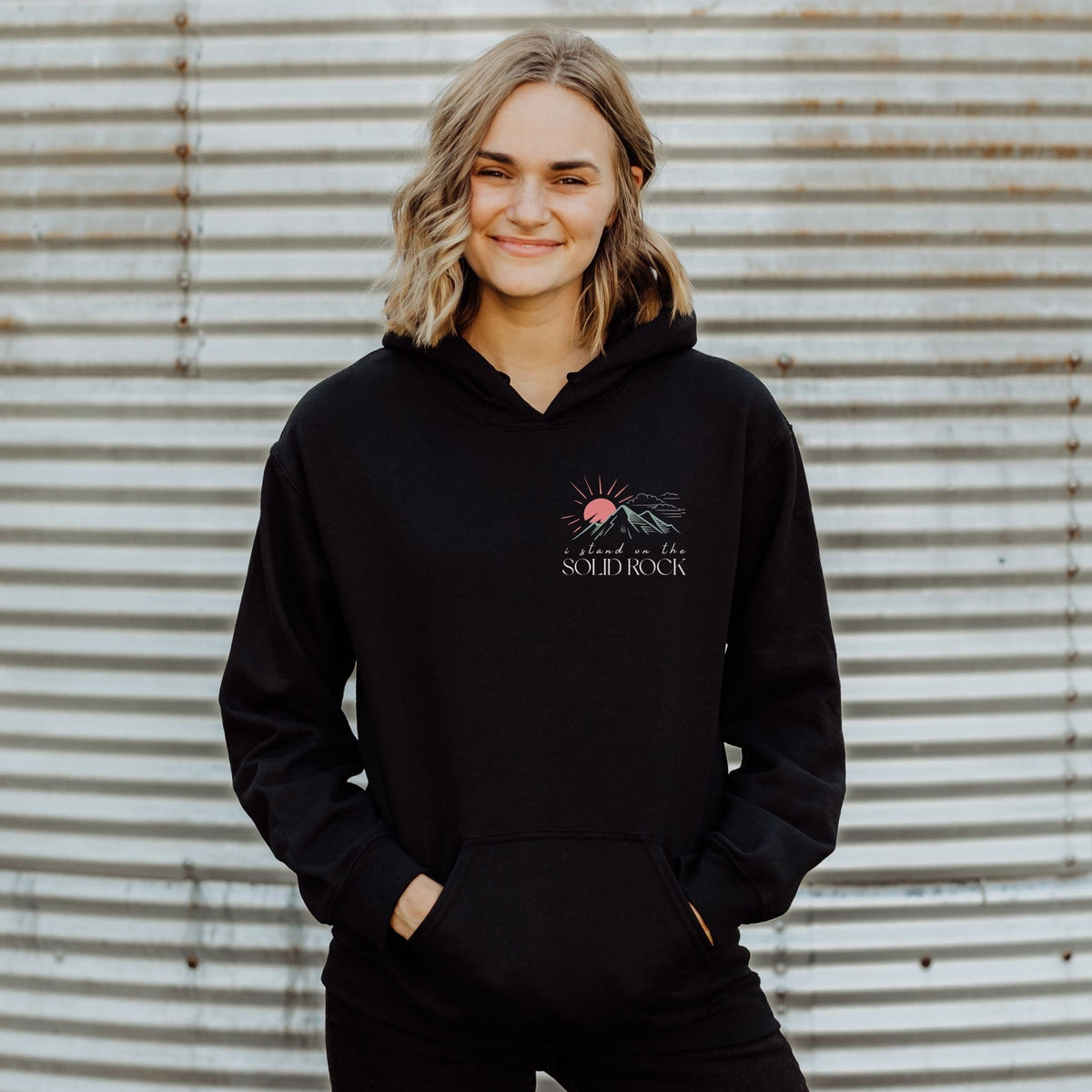 Person wearing a black hoodie with a logo and text, standing against a corrugated metal background