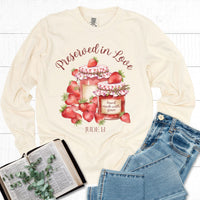 Beige long-sleeve shirt with strawberry-themed design and denim jeans on a white wooden surface.