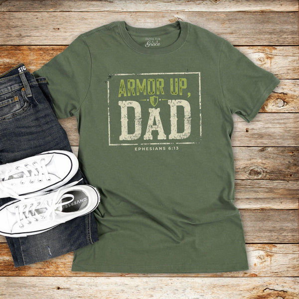Green t-shirt with 'Armor Up, Dad' text on a wooden surface with jeans and sneakers.