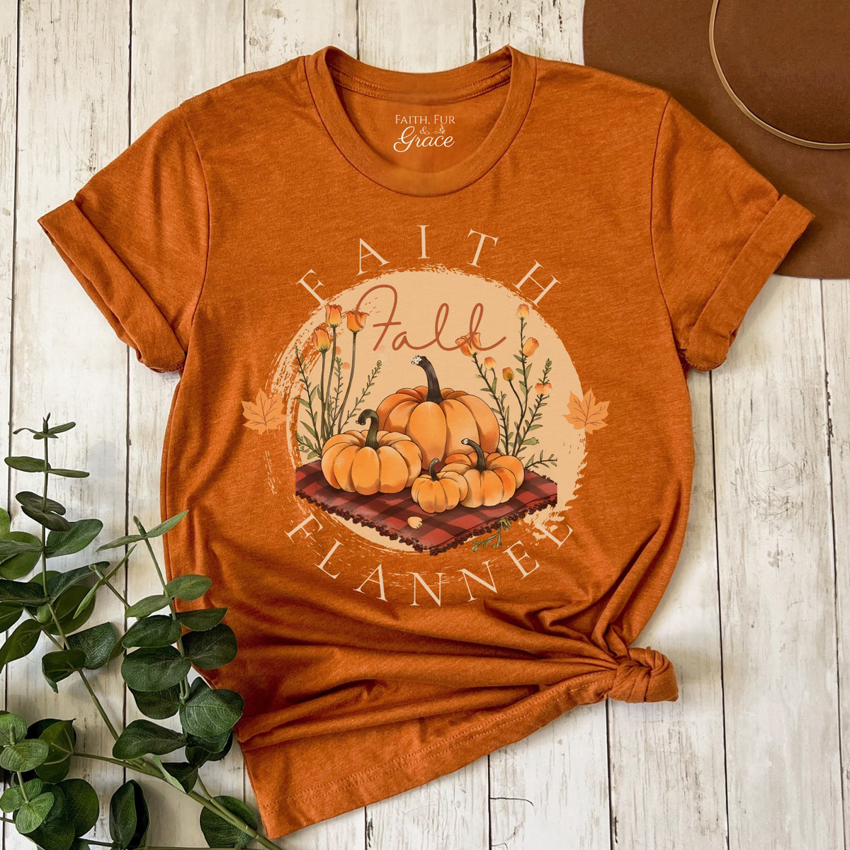 Orange t-shirt with 'Faith Fall Flannel' design on a wooden surface with a plant.