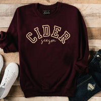 Maroon sweatshirt with 'CIDER season' text on a wooden surface with white sneakers and jeans.