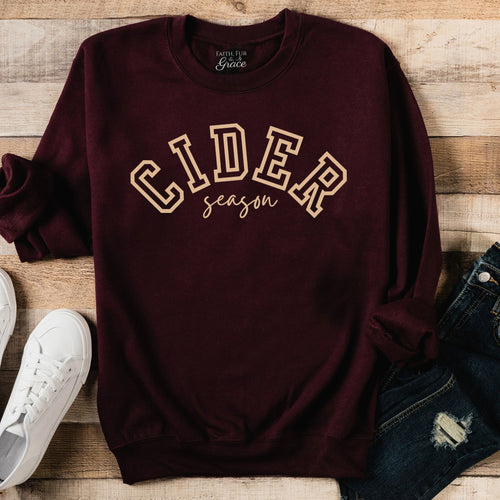 Maroon sweatshirt with 'CIDER season' text on a wooden surface with white sneakers and jeans.
