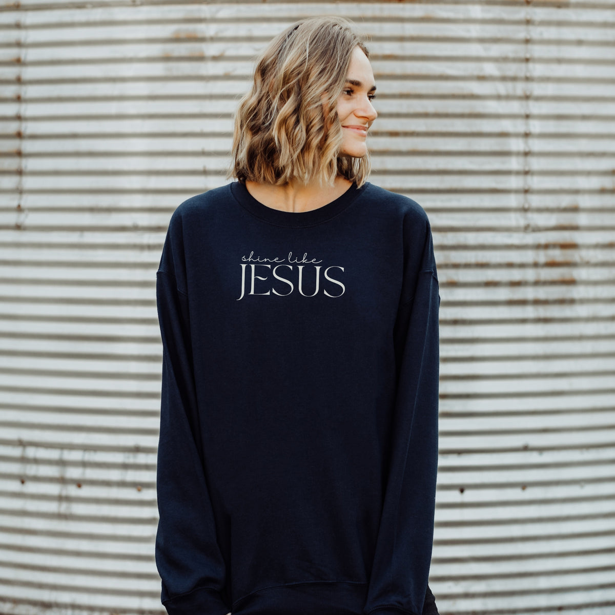 Person wearing a navy sweatshirt with 'shine like Jesus' text against a corrugated metal background