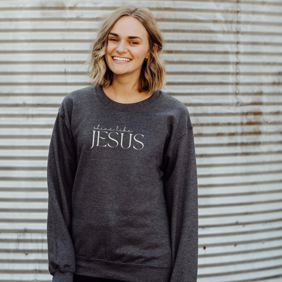 Person wearing a dark gray sweatshirt with 'shine like Jesus' text against a corrugated metal background