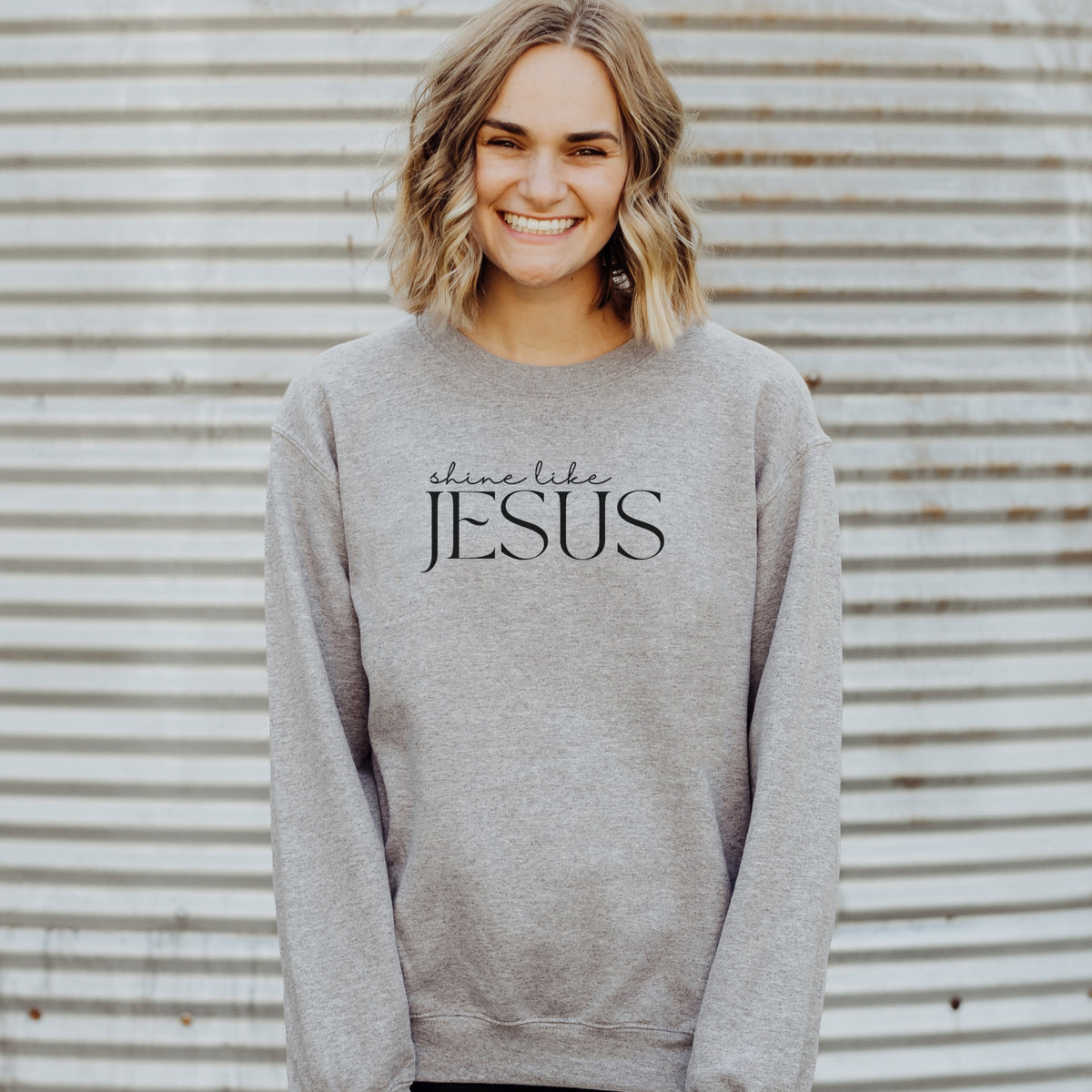 Person wearing a gray sweatshirt with 'shine like JESUS' text against a corrugated metal background