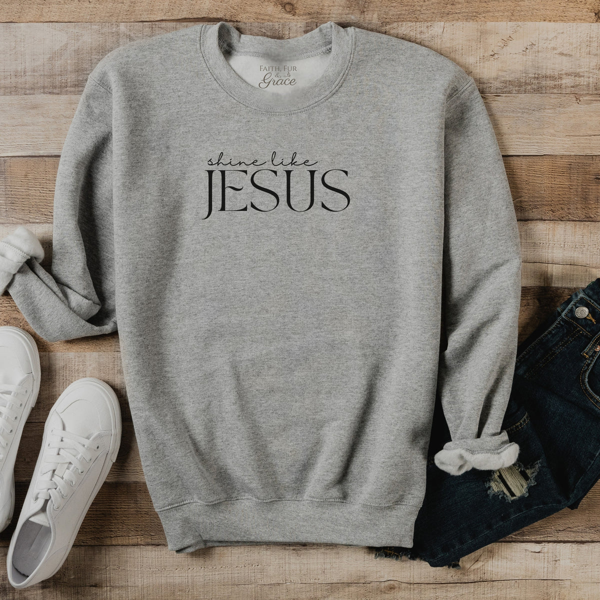 Gray sweatshirt with 'Shine Like Jesus' text on a wooden surface with shoes and jeans.
