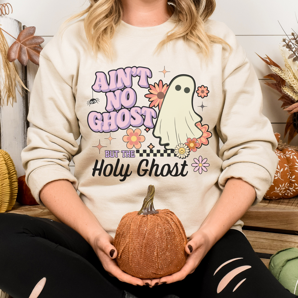 Person wearing a sweatshirt with a ghost graphic and text, holding a small pumpkin.