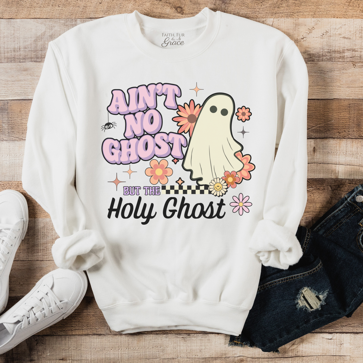 White sweatshirt with ghost graphic and text on a wooden surface with shoes and jeans.