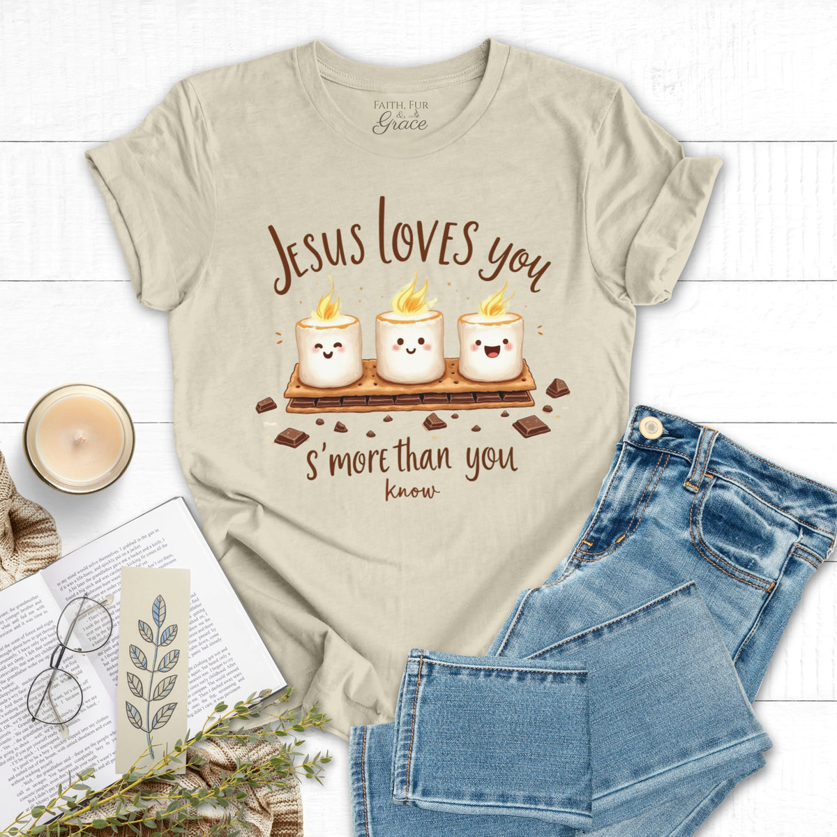 Beige t-shirt with 'Jesus loves you s'more than you know' design, paired with jeans and a candle on a white background.