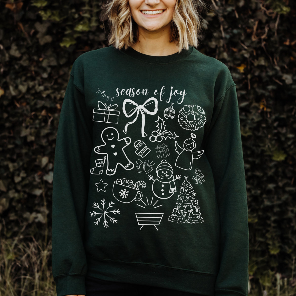 Person wearing a dark green sweatshirt with white Christmas-themed graphics and text 'season of joy' against a natural background.