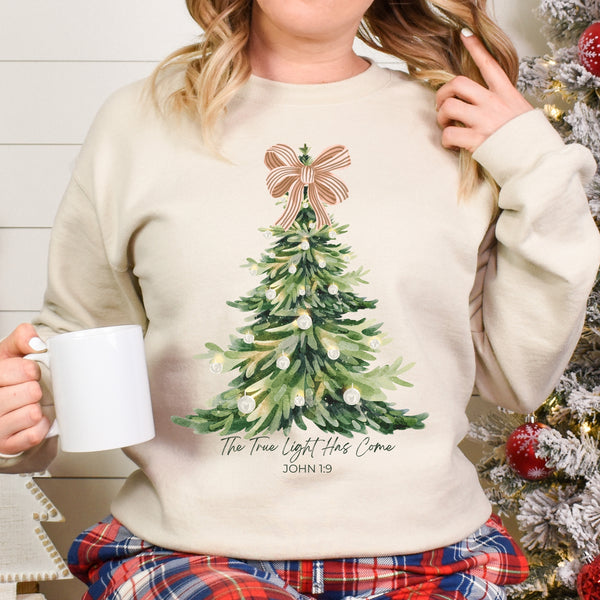 Person wearing a beige sweatshirt with a Christmas tree design, holding a mug, in front of a decorated Christmas tree.