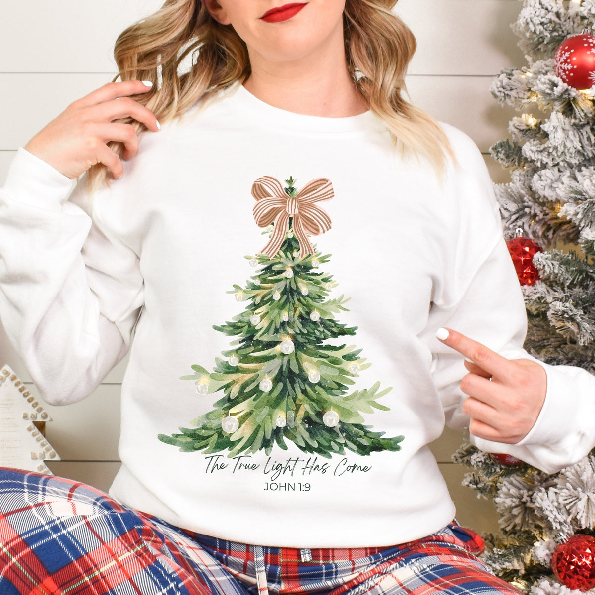 Person wearing a white sweatshirt with a Christmas tree design and plaid pants in front of a decorated Christmas tree.