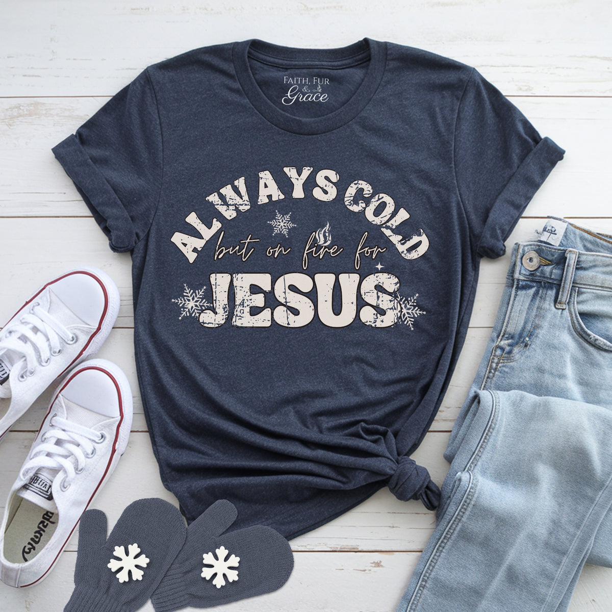 Navy blue t-shirt with 'Always Cold but on Fire for Jesus' text, white sneakers, jeans, and mittens on a light wooden surface.