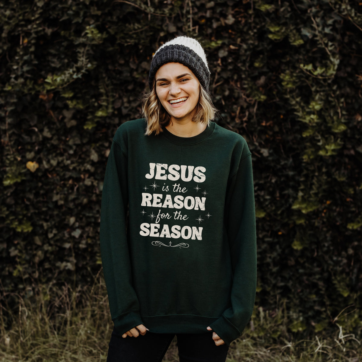 Person wearing a dark green sweatshirt with 'Jesus is the Reason for the Season' text against a forest of trees.