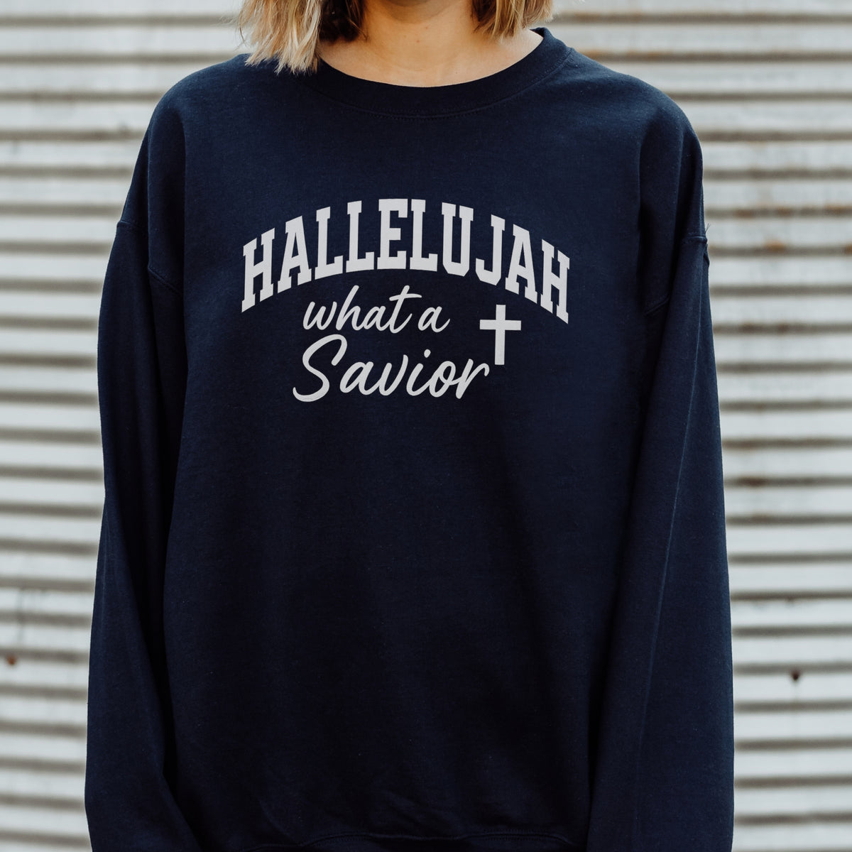 Navy blue sweatshirt with 'HALLELUJAH what a Savior' text and cross symbol on a corrugated metal background