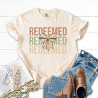Beige t-shirt with 'REDEEMED' text and bow graphic, paired with jeans and boots on a white wooden surface.
