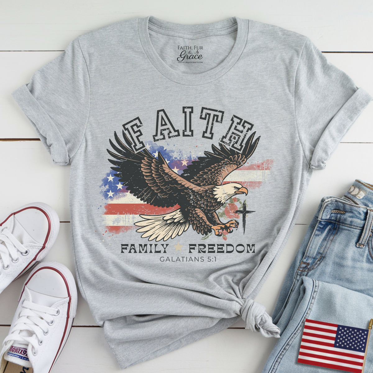 Gray t-shirt with eagle graphic and text on a wooden surface with shoes and jeans.