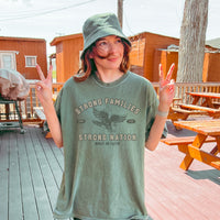 Person wearing a green t-shirt with text and an eagle graphic, standing outdoors on a wooden deck.