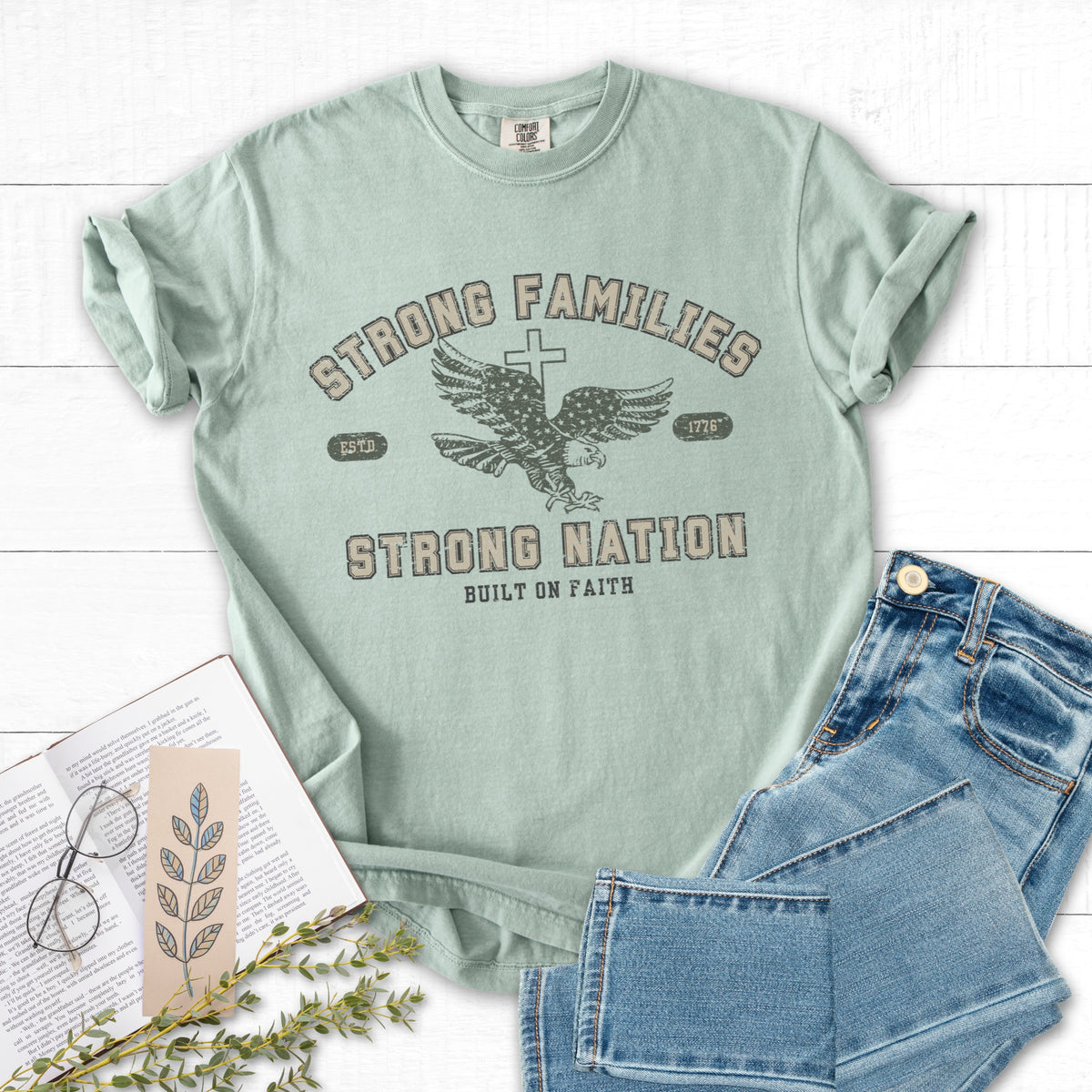 Mint green t-shirt with graphic design and text, paired with jeans and a book on a white surface.