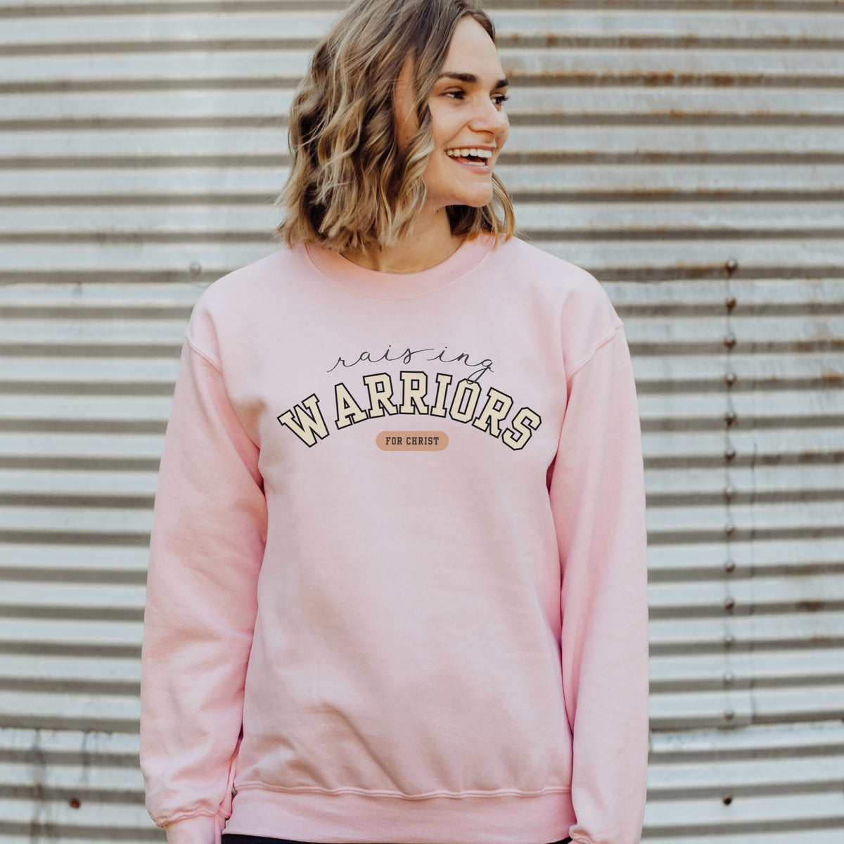 Woman wearing a pink sweatshirt with 'Raising Warriors for Christ' text against a corrugated metal background